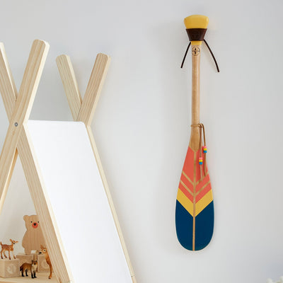 Pagaie décorative en bois peinte de trois couleurs. Bleu, rose et jaune. Pagaie de petite taille fixée è un mur blanc à l'aide d'une attache en cuir de vache huilé – Onquata à Wendake